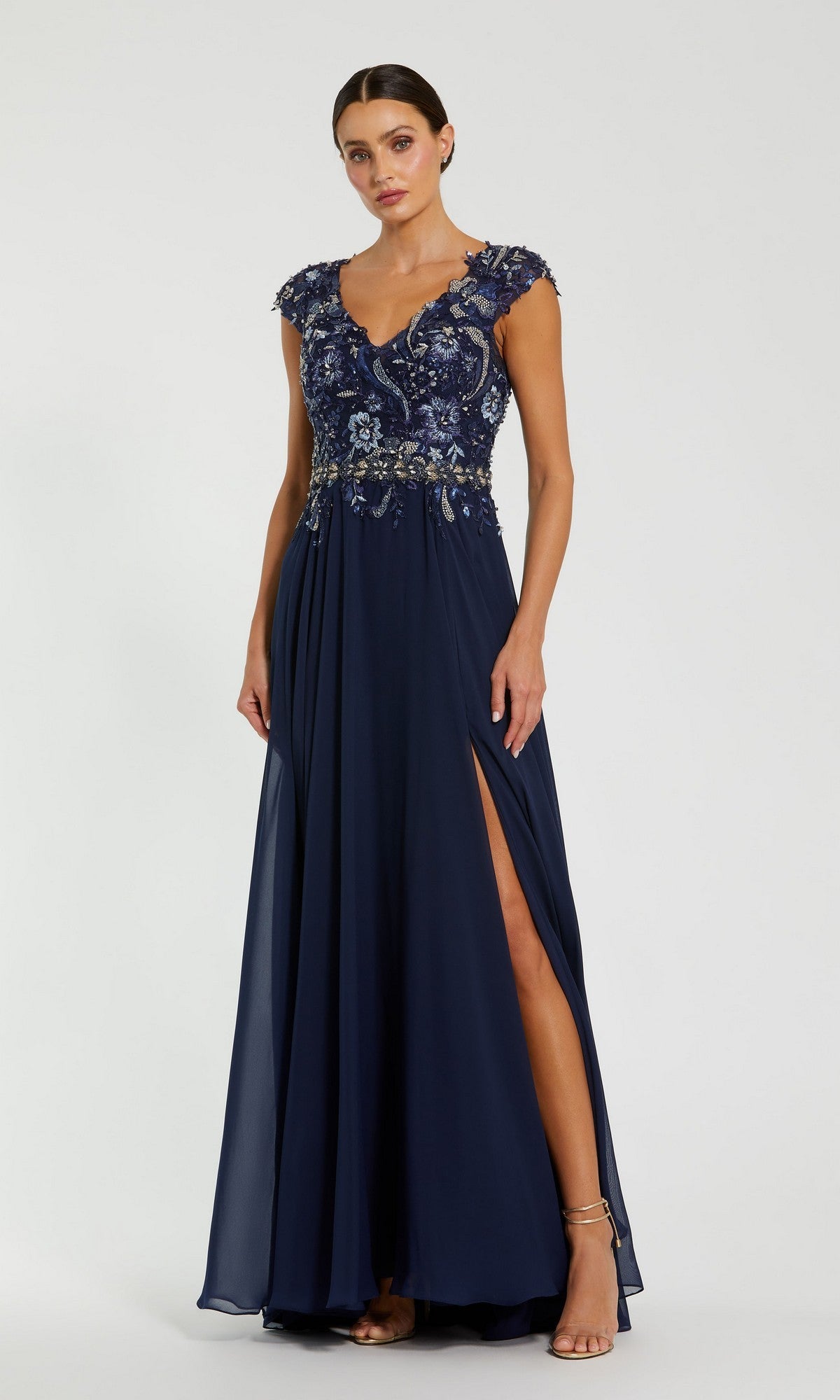 A woman models the Mac Duggal 79365 Cap-Sleeve Long Formal Dress, a navy gown with floral beaded bodice, thigh-high slit, and elegant floor-length flowy silhouette against a plain background.